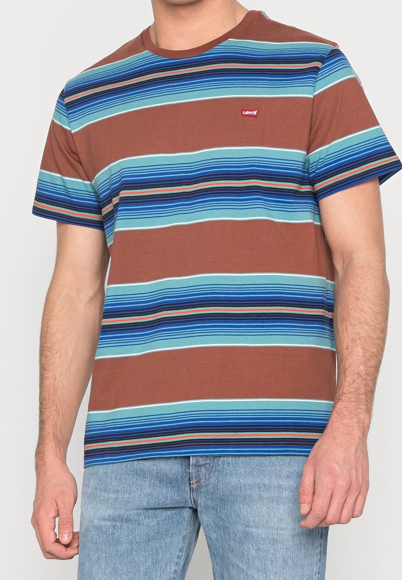 Homme portant un t-shirt marron à manches courtes avec des rayures horizontales bleues et turquoise et un jean bleu clair.