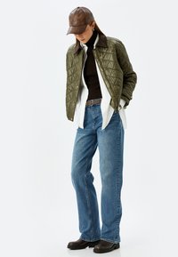 Chaqueta bomber acolchada de color verde oliva con cuello marrón, superpuesta sobre un suéter negro, una camisa blanca y unos vaqueros azules, combinada con botas marrones.