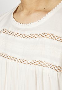Primo piano di un colletto di una blusa bianca con bordo scallop e dettagli in pizzo orizzontale su pelle chiara.