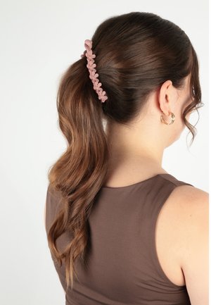 Donna con lunghi capelli castani raccolti in una coda di cavallo fissata da una molletta per capelli floreale rosa, che indossa orecchini a cerchio dorati e una canotta marrone senza maniche.