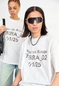 Fehér, túlméretezett pólók merész nyomattal. Az egyik modellen fekete napszemüveg van. A háttér egyszínű. A szöveg tartalmazza: "atelier de Roupa TERRA 02 01/25."