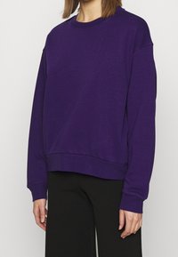 Sudadera de algodón morada con cuello redondo, hombros caídos y corte relajado, combinada con pantalones de pierna ancha negros.