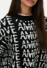 Donna che indossa un maglione nero con parole bianche lavorate a maglia "LOVE" e "AMOUR" ripetute in un motivo, che mostra il colletto e i capelli lunghi.