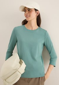 T-shirt à manches longues côtelé couleur sarcelle avec un col rond, associé à un sac beige clair. La personne porte une casquette de couleur claire, se tenant devant un fond neutre.