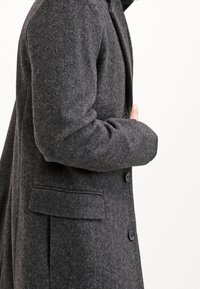Šedý texturovaný kabát s přizpůsobeným střihem, s klopou, dvěma knoflíky a bočními kapsami, vystavený z bočního úhlu.