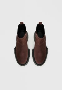 O pereche de cizme slip-on din suede maro, cu tălpi chunky negre, panouri elastice pe lateral și o bandă de prindere în spate, prezentate din perspectiva de sus pe un fundal alb.