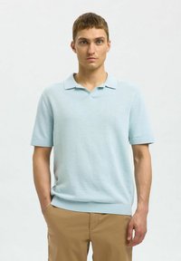 Svijetloplava kratkoslavljena polo majica s klasičnim ovratnikom, rebrastim manžetama i ravnim rubom. U kombinaciji s bež hlačama.