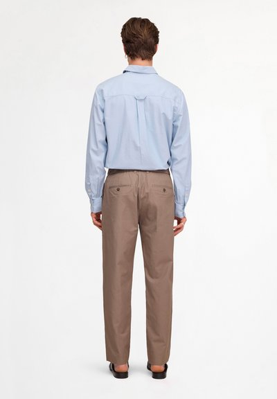 Adulte portant une chemise bleu clair à manches longues et un pantalon marron, debout, dos face à un fond blanc.