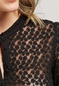 Tissu noir transparent avec un motif de broderie florale, présentant une bordure texturée et des boutons ronds à l'avant. Design délicat et complexe.