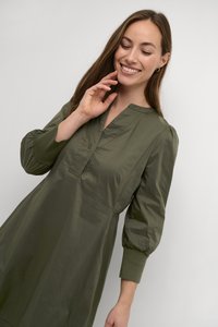 Olivnozeleni outfit iz lahkega materiala, ki ima V-izrez, gumbne detajle in nabran rokav z prilegajočimi manšetami. Gladka tekstura.