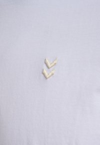 Twee kleine crème kleurige geborduurde chevrons die naar beneden wijzen op een witte stofachtergrond.