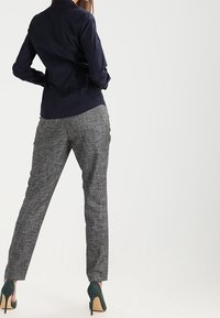 Marineblauw overhemd met lange mouwen, gecombineerd met grijs geruite broeken en donkergroene hakken. Focus op de textuur en het slanke model van de outfit.