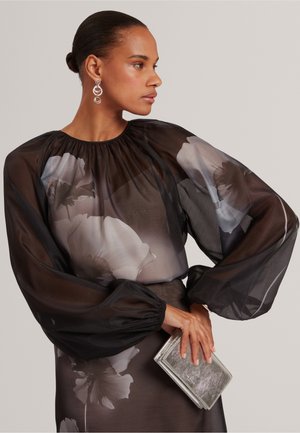 Kvinde med hår sat i knold iført en gennemsigtig sort blomstret bluse og holder en sølv clutch, set fra siden mod en enkel baggrund.