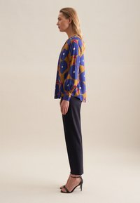 Blusa blu e gialla con motivi a cerchi e floreali, abbinata a pantaloni neri aderenti e tacchi a strappo neri, vista di lato.