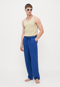 Beige tank top og blå linbukser med snor i livet. Personen har på seg hvite, vinklete solbriller og er barføtt.