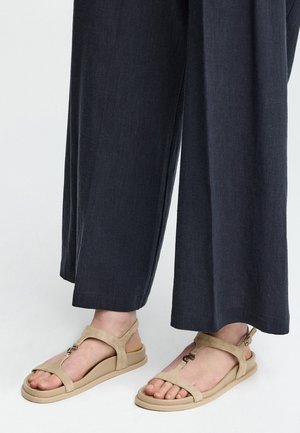 Sandales plates beiges avec des détails de bride et de boucle portées avec un pantalon large gris foncé ample.