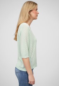 Pull léger vert pâle en tricot, avec une coupe décontractée, des manches courtes et un motif texturé comportant des lignes diagonales et de petites ouvertures.
