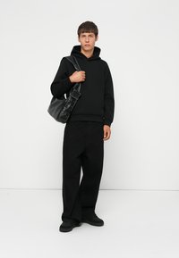 Haut à capuche noir avec une poche avant, associé à un large pantalon noir. Le mannequin tient un sac noir texturé avec des fermetures à pression. Des chaussures noires complètent le look.