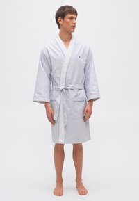 Νέος άνδρας όρθιος με γυμνά πόδια, φορώντας μπλε και λευκή ριγωτή ρόμπα μήκους μέχρι το γόνατο με ζώνη δεμένη στη μέση, κοιτάζει προς τα πλάγια.