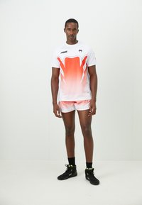 Man standing in white and orange sportswear with black high-top sneakers against a plain white background.