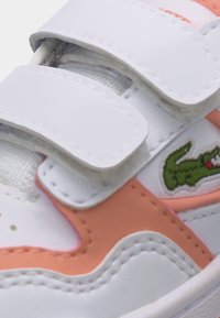 Άσπρο και ροζ αθλητικό παπούτσι με διπλά λουράκια βέλκρο και μικρό πράσινο λογότυπο κροκόδειλου στην πλαϊνή πλευρά.