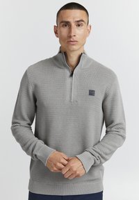 Lys grå genser med kvart glidelås, som har vertikal ribbet tekstur og et lite logo-patch på brystet. Slim-fit design.