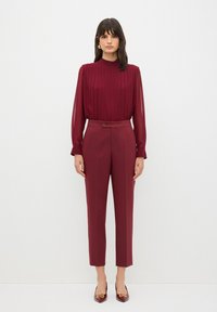 Blusa plissettata bordeaux con collo alto, abbinata a pantaloni su misura coordinati con dettagli a bottone e gambe slim dritto.