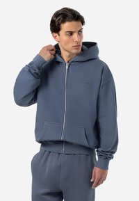 Mikina s kapucňou a zipsom v jemnej modrej farbe, s klokankou a rebrovanými manžetami. Obsahuje jemný detail loga na hrudi.