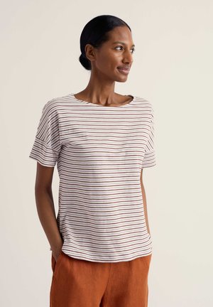 Tumejate juustega naine, kellel on sooritusmustriga valge triibuline t-särk ja roostepunased püksid, seisab käed taskutes, vaadates paremale.