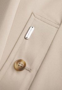 Blazer in tessuto beige con una tasca visibile, dotato di un bottone rotondo marrone e un'etichetta argentata con la scritta "QUINTESSENCE". Texture liscia.