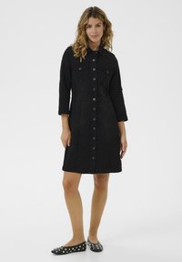 Vestido camisa de denim negro con un cierre de botones en la parte delantera, cuello puntiagudo, dos bolsillos en el pecho y mangas de tres cuartos. Combinado con zapatos planos negros.