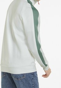 Hvid zip-up jakke med grønne sidelister, høj krave og ribbede manchetter. Blød tekstur med en afslappet pasform, parret med blå jeans.