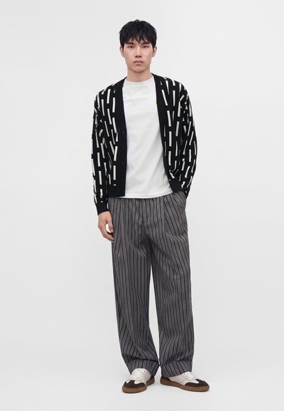 Homme portant un cardigan à motif noir et blanc, un t-shirt blanc, un pantalon large gris à fines rayures et des chaussures blanches et marron, debout devant un fond blanc.