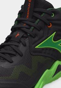 Μαύρο και πράσινο αθλητικό παπούτσι με ζιγκ-ζαγκ μοτίβο, φωτεινό πράσινο λογότυπο, πράσινη σόλα, μαύρα κορδόνια και πορτοκαλί λεπτομέρεια στη γλώσσα και το μεταλλικό κρίκο δέσης.