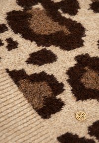 Plaid en laine présentant une base beige avec un grand motif léopard marron foncé et marron clair. Comprend un petit détail de logo doré.