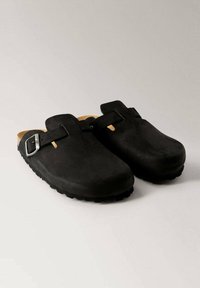 Black suede clogs with a rounded toe, featuring a single adjustable buckle strap and a cushioned tan insole. Treaded rubber sole.