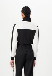 Veste de sport noir et blanc avec un col haut, des manches longues et une taille élastique. Tissu lisse ; panneaux contrastants aux épaules et sur les manches.