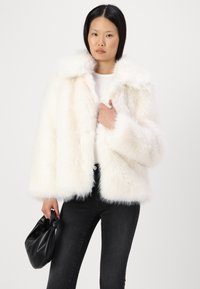 Giacca in ecopelle bianca con ampio colletto e texture morbida, caratterizzata da maniche lunghe e una forma boxy, indossata sopra un top aderente e jeans scuri.