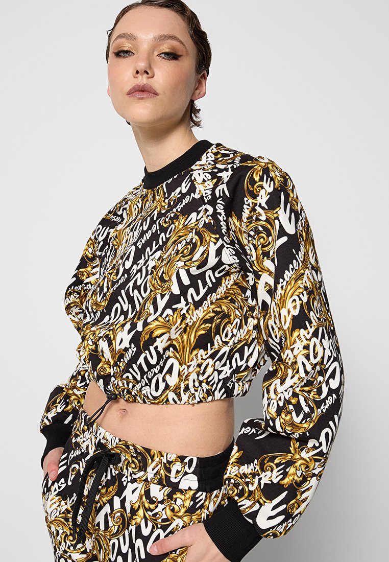 Schwarzes verkürztes Sweatshirt mit auffälligen weißen und goldenen Mustern, featuring einem runden Halsausschnitt und gerippten Bündchen. Das Muster umfasst Text und verschnörkelte Designs.