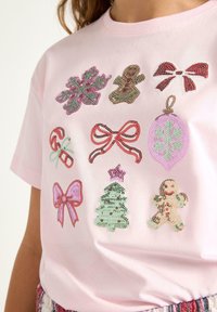 Pink kortærmet T-shirt med dekorerede, syede jule-symboler: snefnug, slikstokke, sløjfer, peberkager, en julekugle og et juletræ.