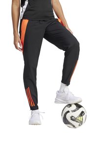 Schwarze Sporthosen mit orangefarbenen Seitenstreifen, enganliegendes Design. Weiße Sneakers getragen, Fuß ruhend auf einem schwarz-weißen Fußball mit Logo.