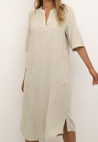 Vestido a rayas en beige y blanco, de longitud hasta la rodilla con un escote sin collar, mangas cortas y aberturas laterales para facilitar el movimiento.