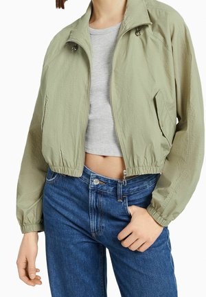 Woman wearing light green zip-up cropped jacket over gray ribbed crop top and blue high-waisted jeans, hand in pocket.