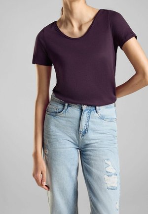 Woman wearing a fitted dark purple short-sleeve top and light blue high-waisted ripped jeans against a plain gray background.