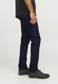 Dunkelblaue Jeans aus Denim mit geradem Schnitt, dezenten Nähten und einem Detail an der Gesäßtasche. Getragen mit schwarzen eleganten Schuhen.