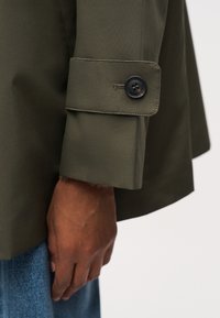 Μανίκι σκούρου ελαιώδους πράσινου μπουφάν με λουράκι και κουμπί, φορεμένο πάνω από το χέρι ενός ατόμου και μπλε τζιν παντελόνι.