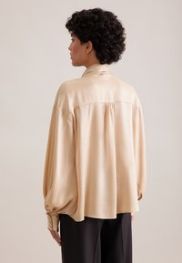 Blusa di satin beige chiaro con maniche lunghe e voluminose, polsini con bottoni, colletto alto e design con schiena arricciata. Abbinata a pantaloni scuri.