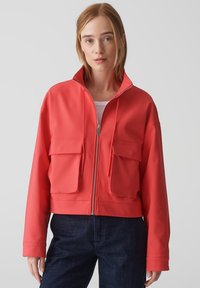 Korallenfarbene, kurz geschnittene Jacke aus glattem Material mit einem hohen Kragen, zwei großen Fronttaschen und einem durchgehenden Reißverschluss.