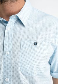 Světle modrá košile s krátkým rukávem, s puntíkovaným vzorem, s límečkem na knoflíky a malou náprsní kapsou s detailem knoflíku značky.