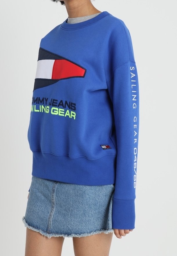 Sweat-shirt bleu avec un logo central vibrant en rouge, blanc et noir. Affiche "SAILING GEAR" en vert néon sur le devant et la manche. Texture douce.